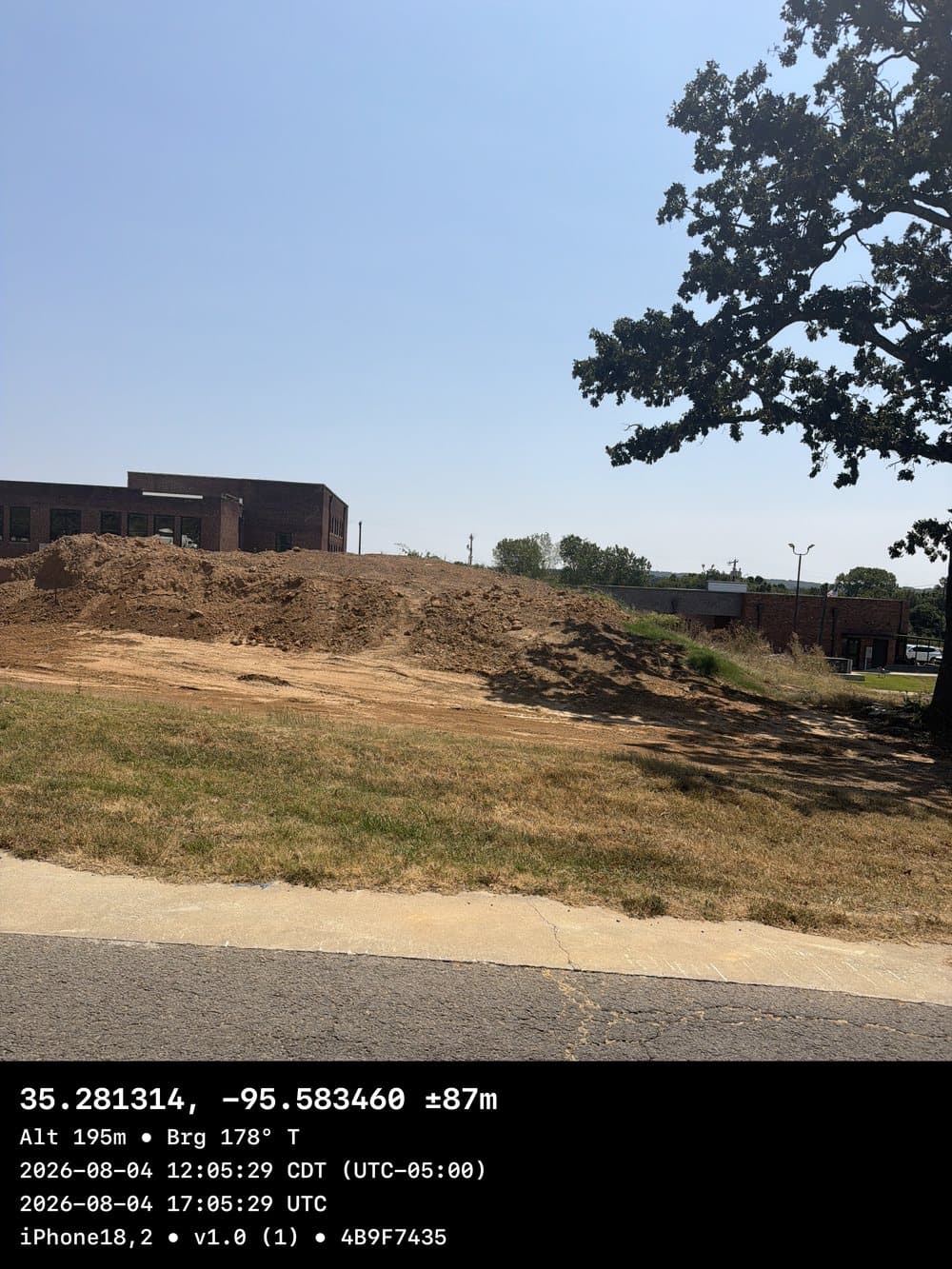 A photo taken with CYA Snapper of a construction site with excavated dirt piles beside a brick building, with a black stamp burned across the bottom reading the coordinates, accuracy of plus or minus 87 meters, altitude, bearing, local and UTC timestamps, device and app version
